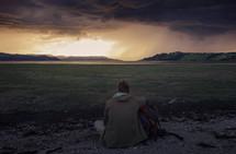 man with a backpack looking out over a grassy area along a shore at sunset 