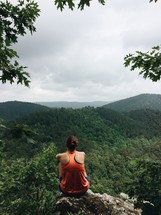 a woman sitting on a rock overlooking a mountain forest 