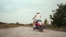 Woman In Helmet Rides On Retro Moped Along Country Road In Summer At Sunset