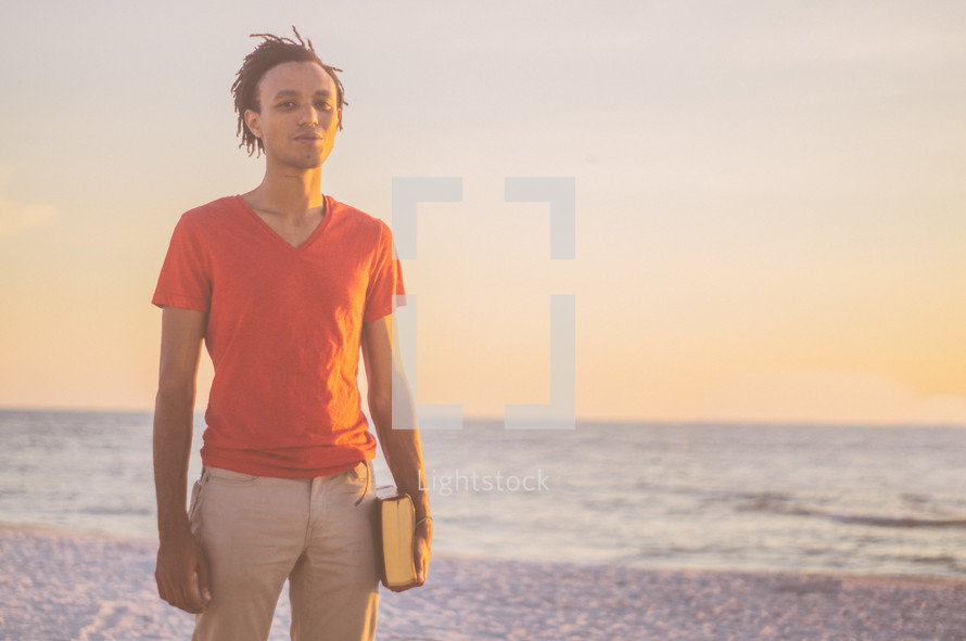 African-American man standing on a beach at sunset holding a Bible 