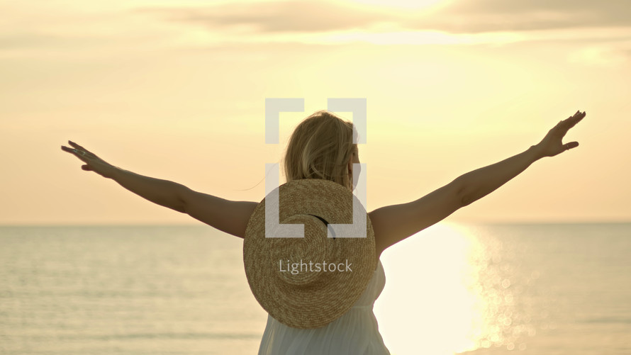 Woman in white dress and straw hat standing with open arms on sea beach. Beauty, nature, travel, hope, harmony concept. High quality photo