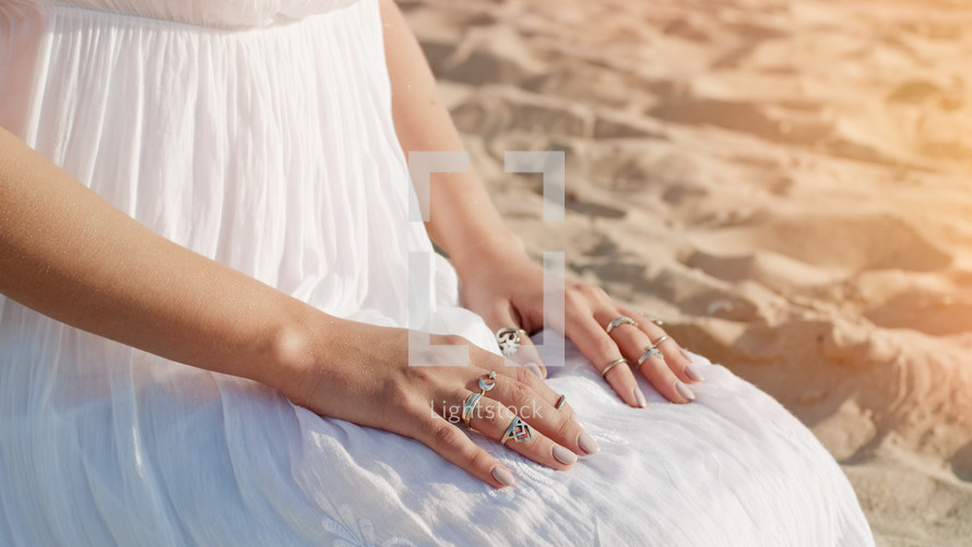 Woman in white dress and boho rings on hands sits on sandy beach alone. Meditation, immersion in inner world, unity with nature. High quality photo