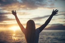 woman standing on a beach with hands raised in praise 