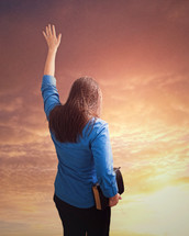 Woman holding a bible with arm raised under and orange sky.