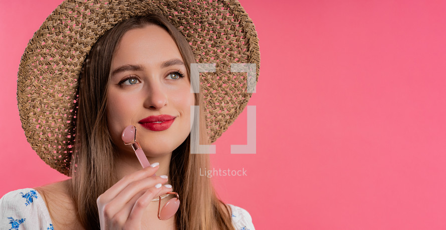 Blonde woman doing face massage with rose quartz stone roller on pink background. Facial self care, beauty rituals, cosmetology, anti aging and anti-wrinkle treatment