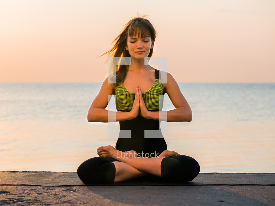 Young caucasian woman in bodysuit relaxing by practicing yoga on the beach near calm sea, close-up of hands, gyan mudra and lotus position. Sunrise background.