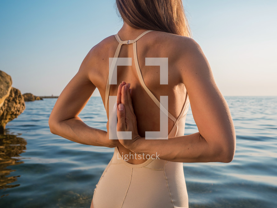 Young caucasian woman in bodysuit relaxing by practicing yoga on the beach near calm sea, close-up of hands, gyan mudra and lotus position. Sunrise background.