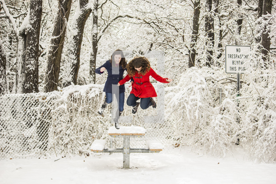 Two girls jumping off a picnic table covered in snow