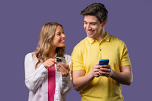 Young couple using mobile device. Woman and man watching, talking, smiling, laughing with smartphone. Social apps, networks, technology concept. Violet studio background.