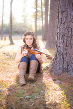 young woman sitting on the forest floor with a violin