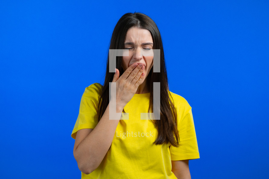 Tired sleepy woman yawns, covers her mouth with hand. Very boring, uninteresting. Blue studio background. High quality photo