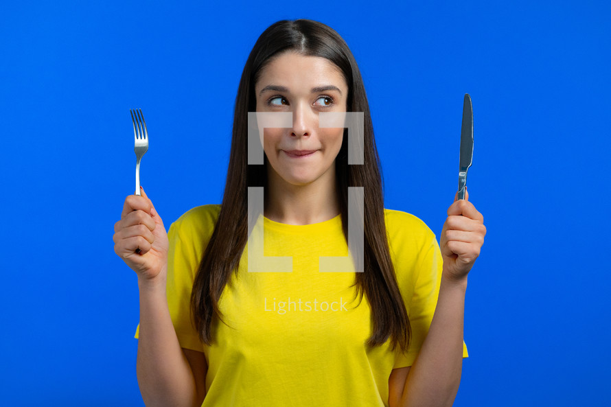 Portrait of hungry woman with fork and knife. Lady with anticipation waiting for serving dinner dishes with cutlery on blue studio background.
