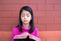 a girl reading a Bible outdoors in front of a brick wall 