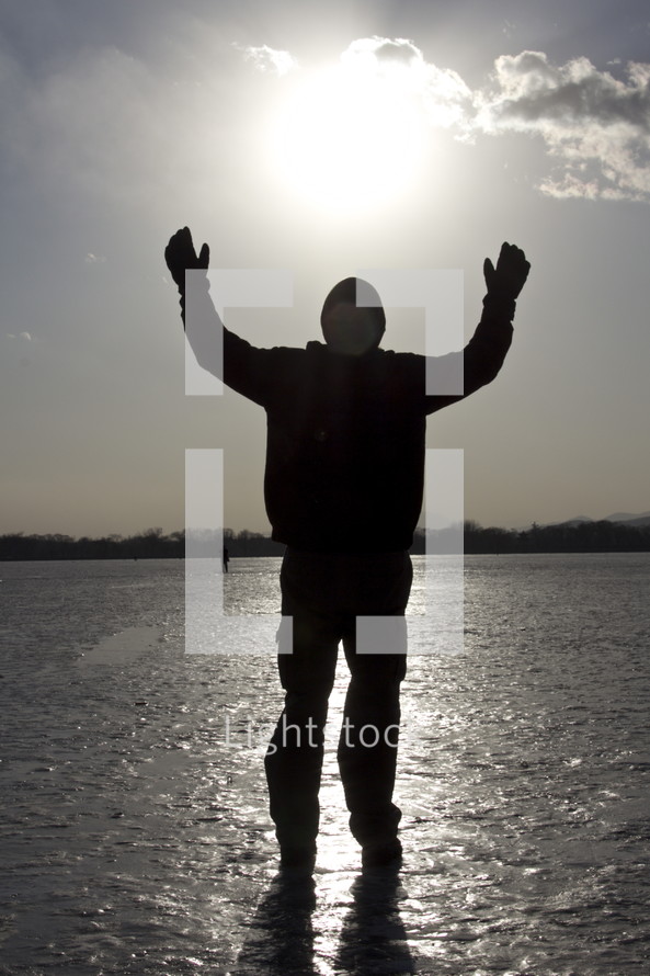 Silhouette of man praising God, hands raised and standing on ice at sunset.
