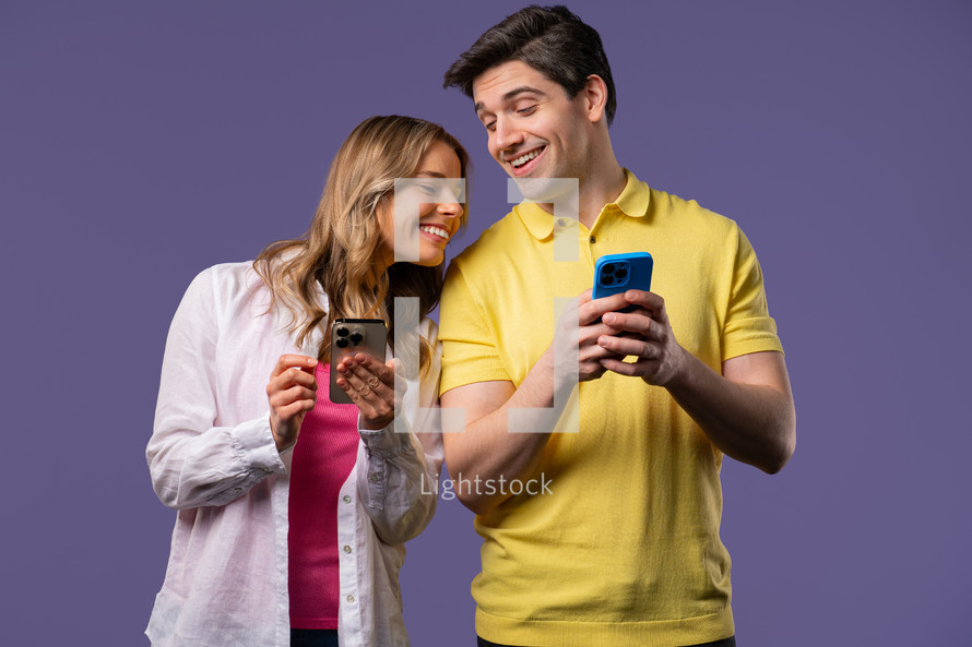 Young couple using mobile device. Woman and man watching, talking, smiling, laughing with smartphone. Social apps, networks, technology concept. Violet studio background.