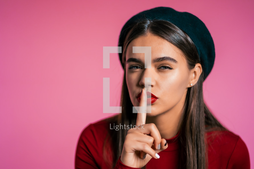 Beautiful woman with red lips holding finger on her lips over pink background. Gesture of shhh, secret, silence. Close up