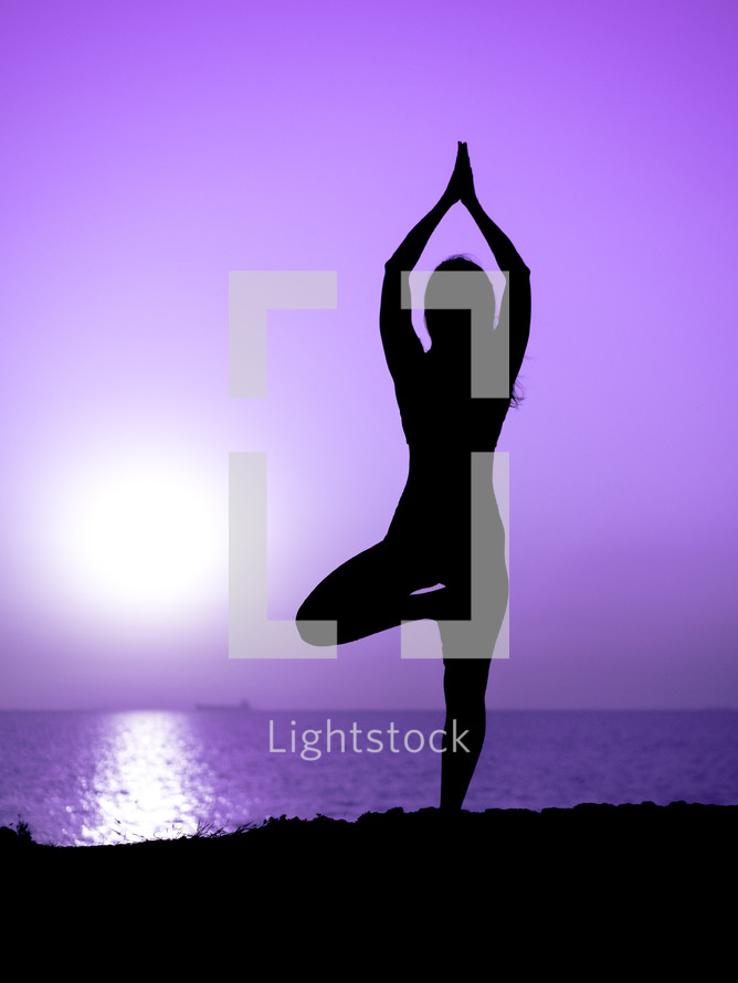 Young slim girl practicing yoga on mountain against ocean or sea at sunrise time. Silhouette of woman in rays of awesome sunset.