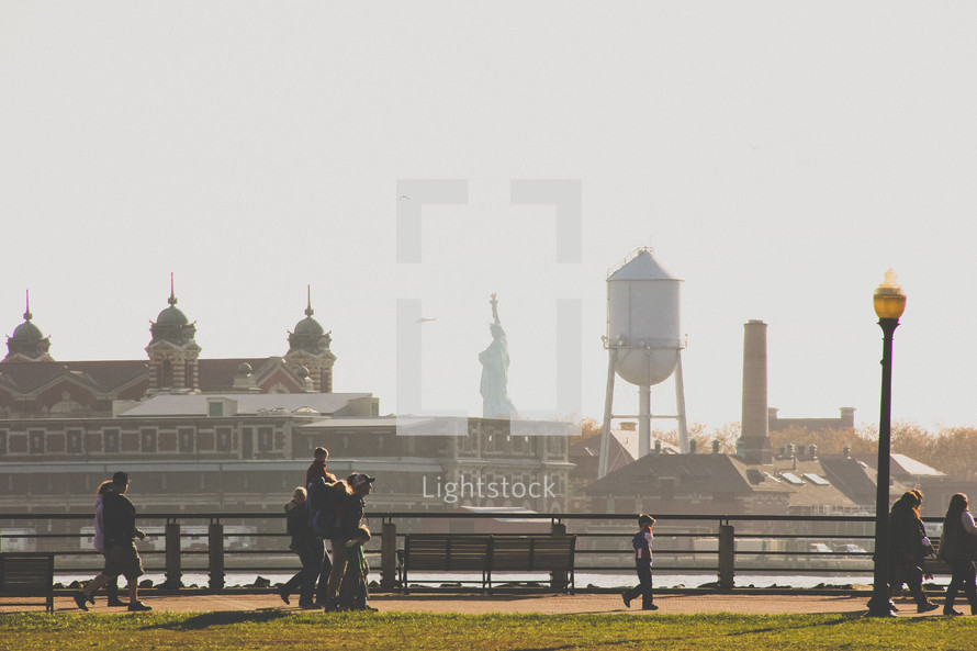 view of the statue of liberty as people walk along a harbor sidewalk 