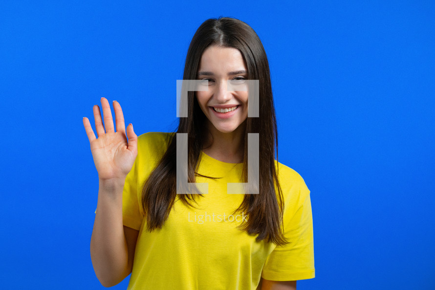 Friendly woman waving hand - goodbye, chao, adios. Parting, say bye to camera. Beautiful girl waving hand on blue studio background. High quality photo