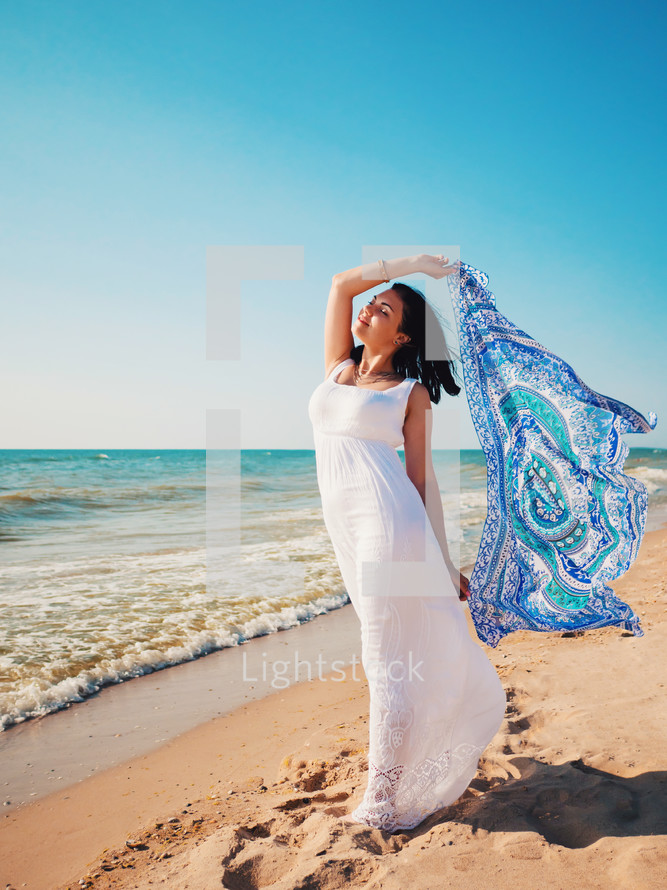 Gypsy young brunette Girl wearing white maxi long dress standing in the sea or ocean with waves and foam. Bohemian clothing style. Boho lifestyle