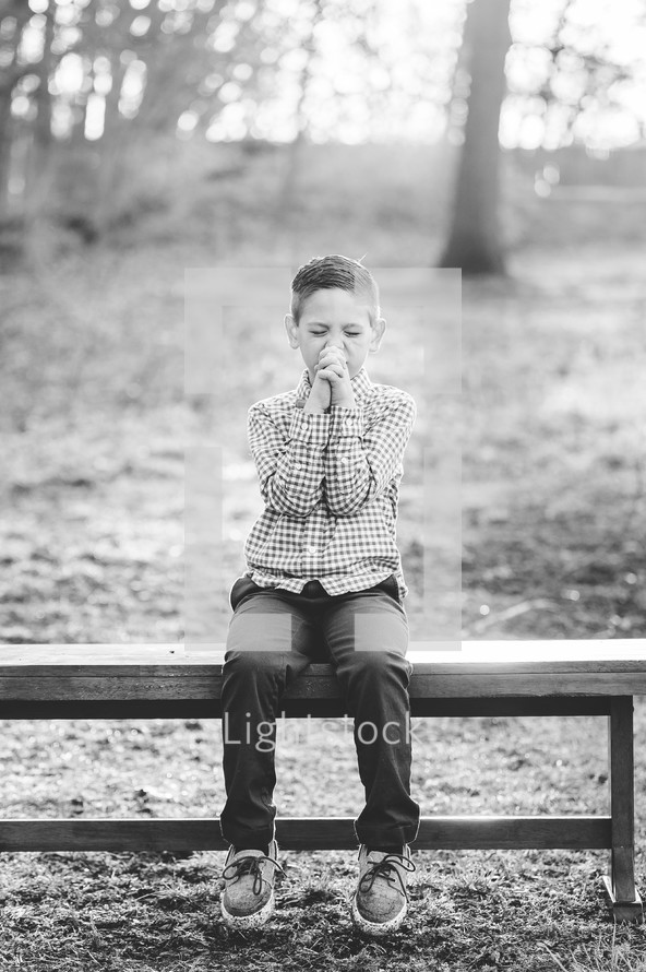 a little boy sitting on a bench outdoors praying 