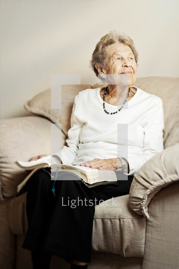 Smiling elderly woman sitting on the couch reading the Bible.