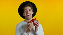 Handsome man holding gift box on yellow studio background and smiles to camera. Happy european hipster guy received present and interested in what's inside.