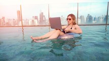 A young entrepreneur woman is working on her computer in by the resort pool in summer time. Beautiful young female model working freelance at swimming pool on laptop.	