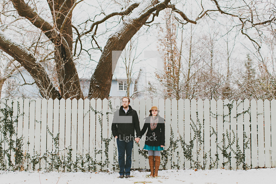 a couple standing in front of a fence in the snow holding each others hands 
