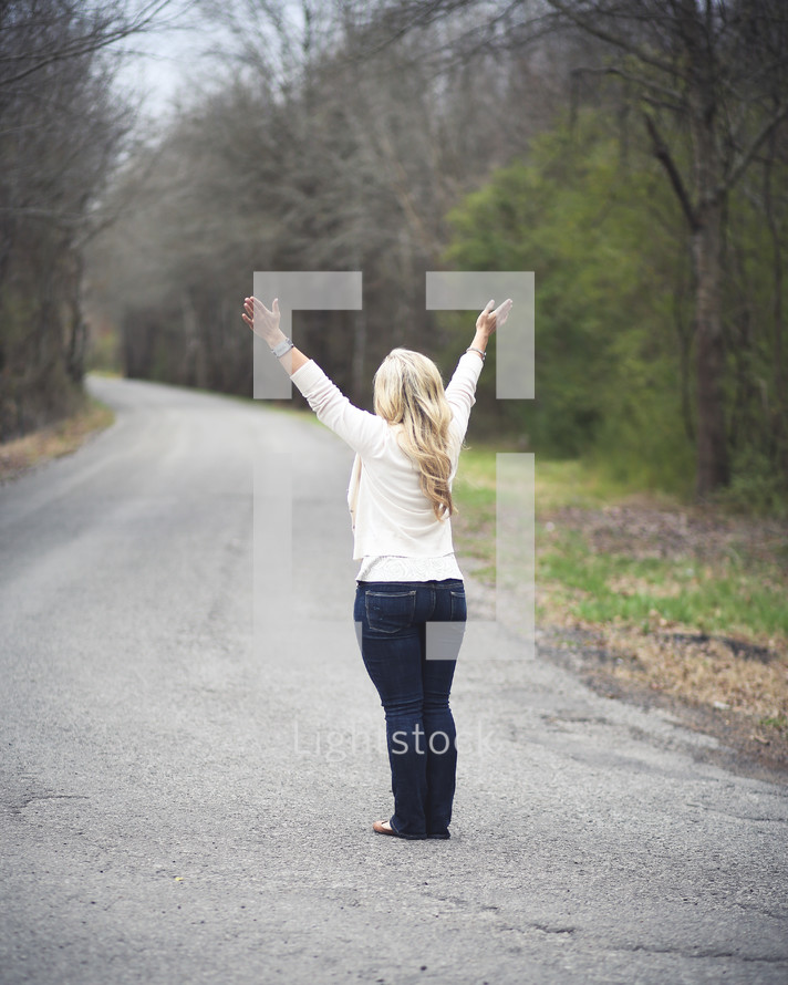 woman standing in the middle of an open road with her arms raised to God