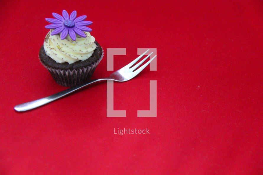 Cupcake decorated with cream cheese, sprinkles and fondant purple flower against a red background. 