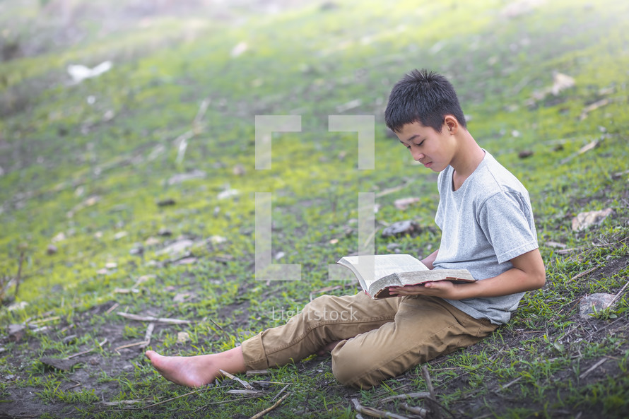 a boy sitting on a hill reading a Bible 