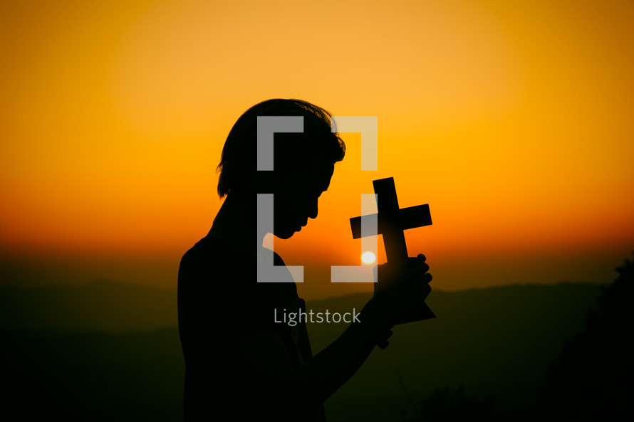 Young man praying and holding Cross at sunset  