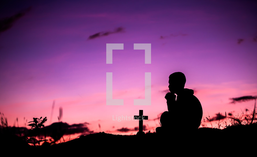 silhouette of a cross and a boy praying outdoors at sunset 