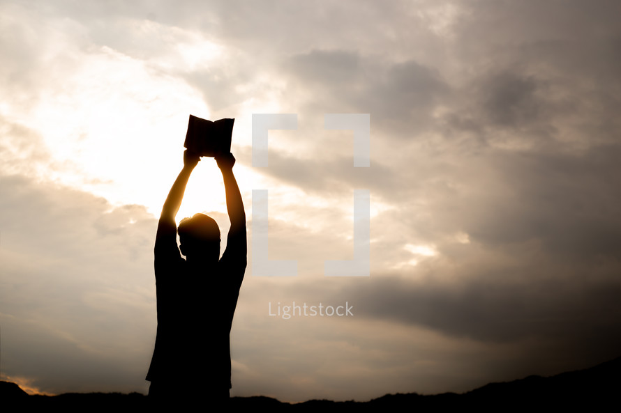 silhouette of a man holding up a Bible outdoors 