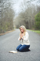 woman kneeling in prayer in the middle of a road next to an open Bible