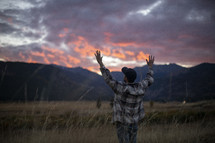 a man with raised hands standing in a field at sunset 