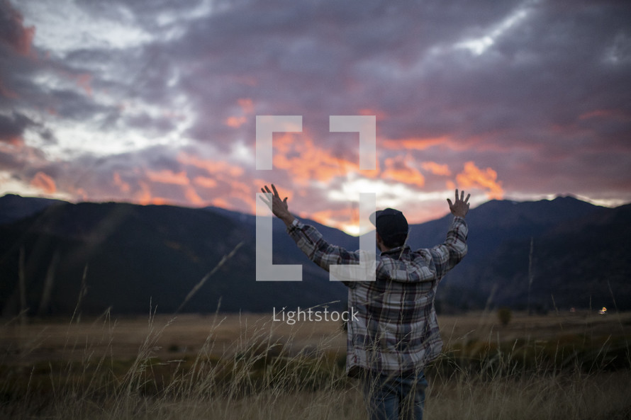a man with raised hands standing in a field at sunset 