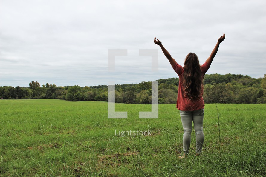 young woman standing in a field with arms raised 