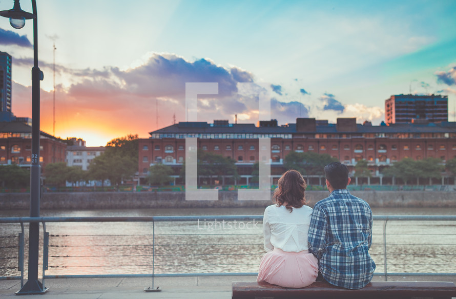 a couple sitting on a bench looking out at a river at sunset 