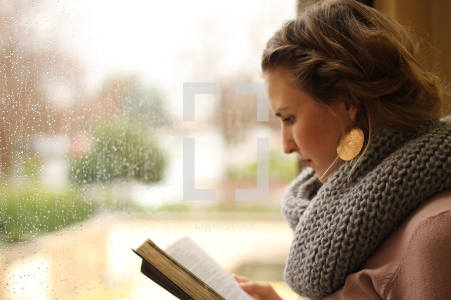 a woman reading a Bible and rain out a window 