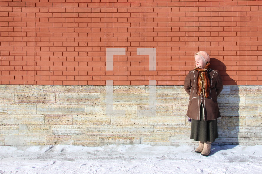 Russian woman leaning against a brick wall, warming herself in the sun