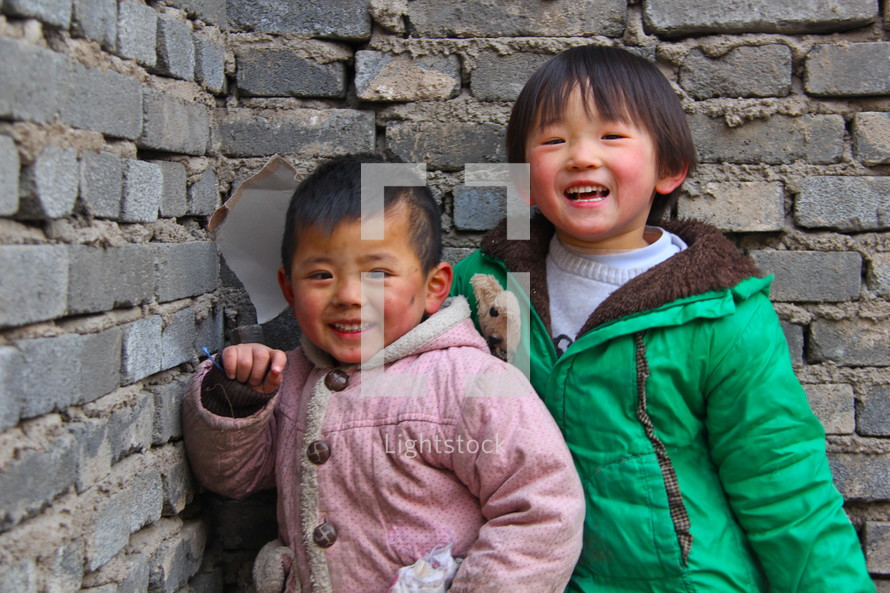 Two Mongolian children smiling standing in a corner of a concrete block building [For more, try search 'Ethnic smile']