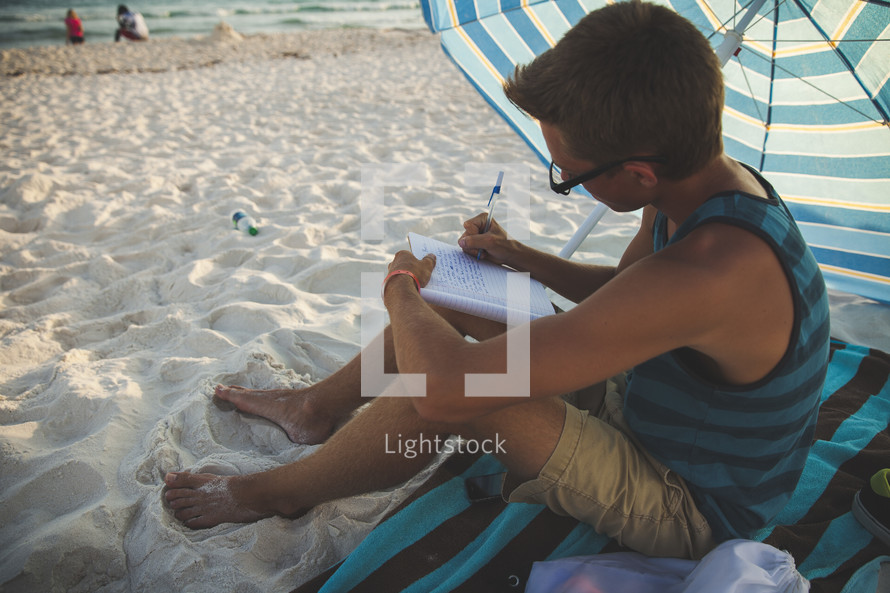 man writing in a journal on the beach under an umbrella 