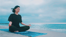  Fitness woman in sports clothing practicing yoga on the sea ocean beach close up to water. Dolly shot. Calm young woman sitting in lotus pose.
