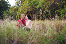 Couple sitting and embracing in a weedy field bordered with trees, shrubs, bushes