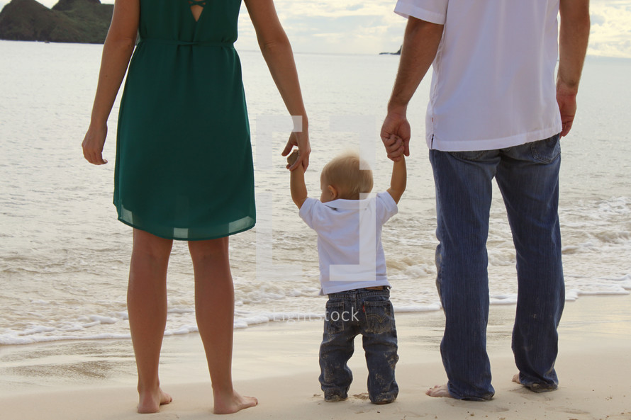 a mother and father holding hands with their toddler son on a beach 