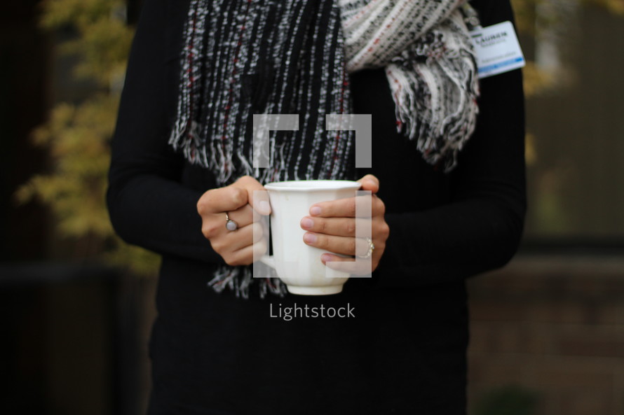 a woman standing outdoors in a scarf holding a mug 