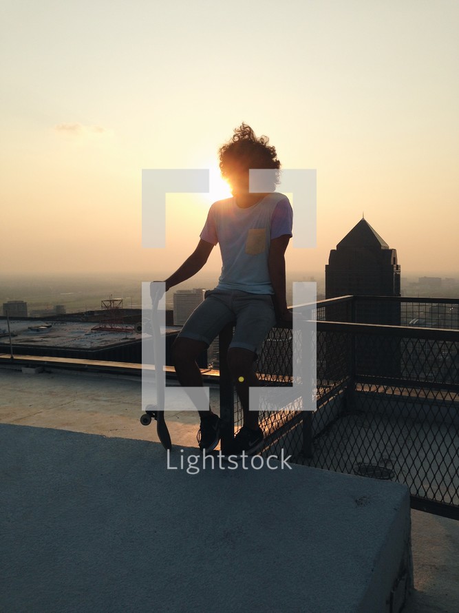 young man with a skateboard sitting on a roof in a city 