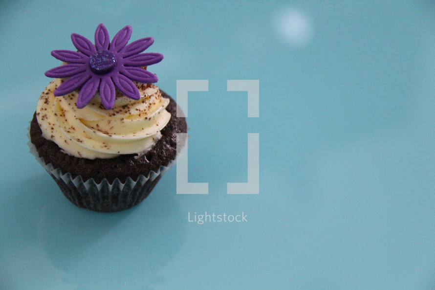 Cupcake decorated with cream cheese, sprinkles and a purple flower against a blue background 